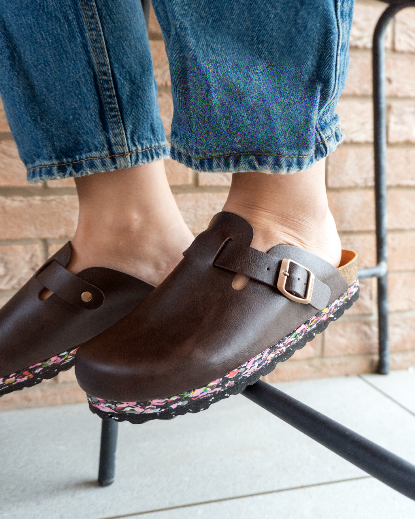 Person wearing Nuoceans Nuos sustainable vegan clogs made from recycled ocean waste, dark brown colour, sitting outdoors against brick wall