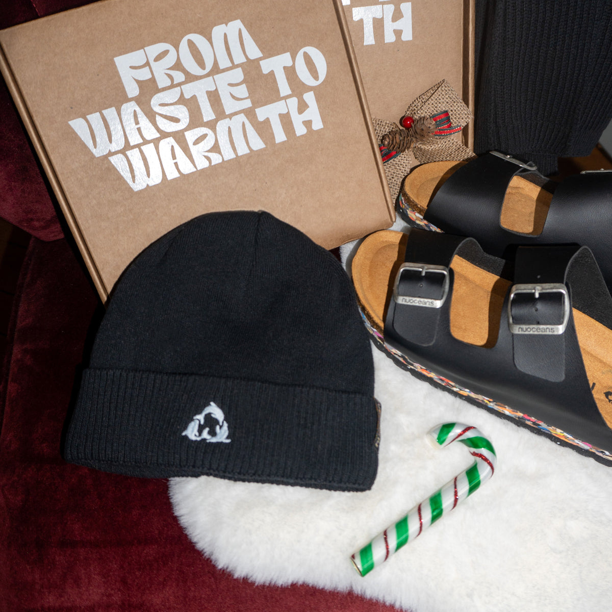 Black beanie, cork sandals, and 'From Waste to Warmth' boxes on a red surface.